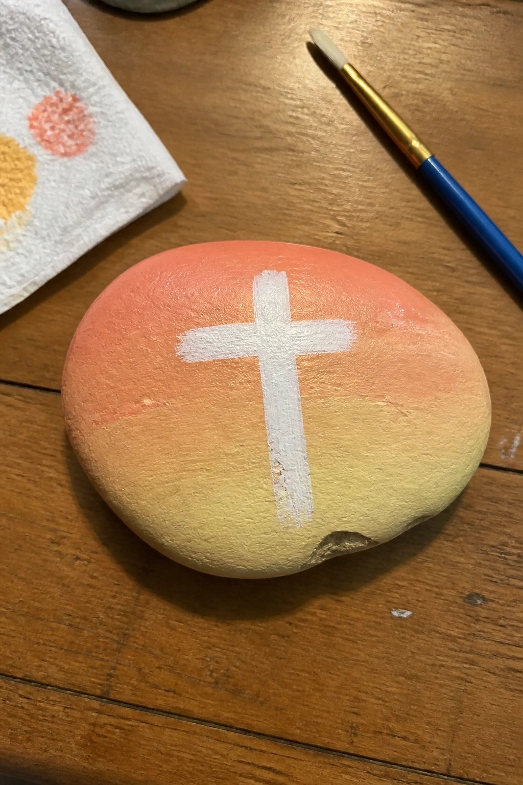 Oval rock with orange-yellow gradient and white cross beside paintbrush on wooden table