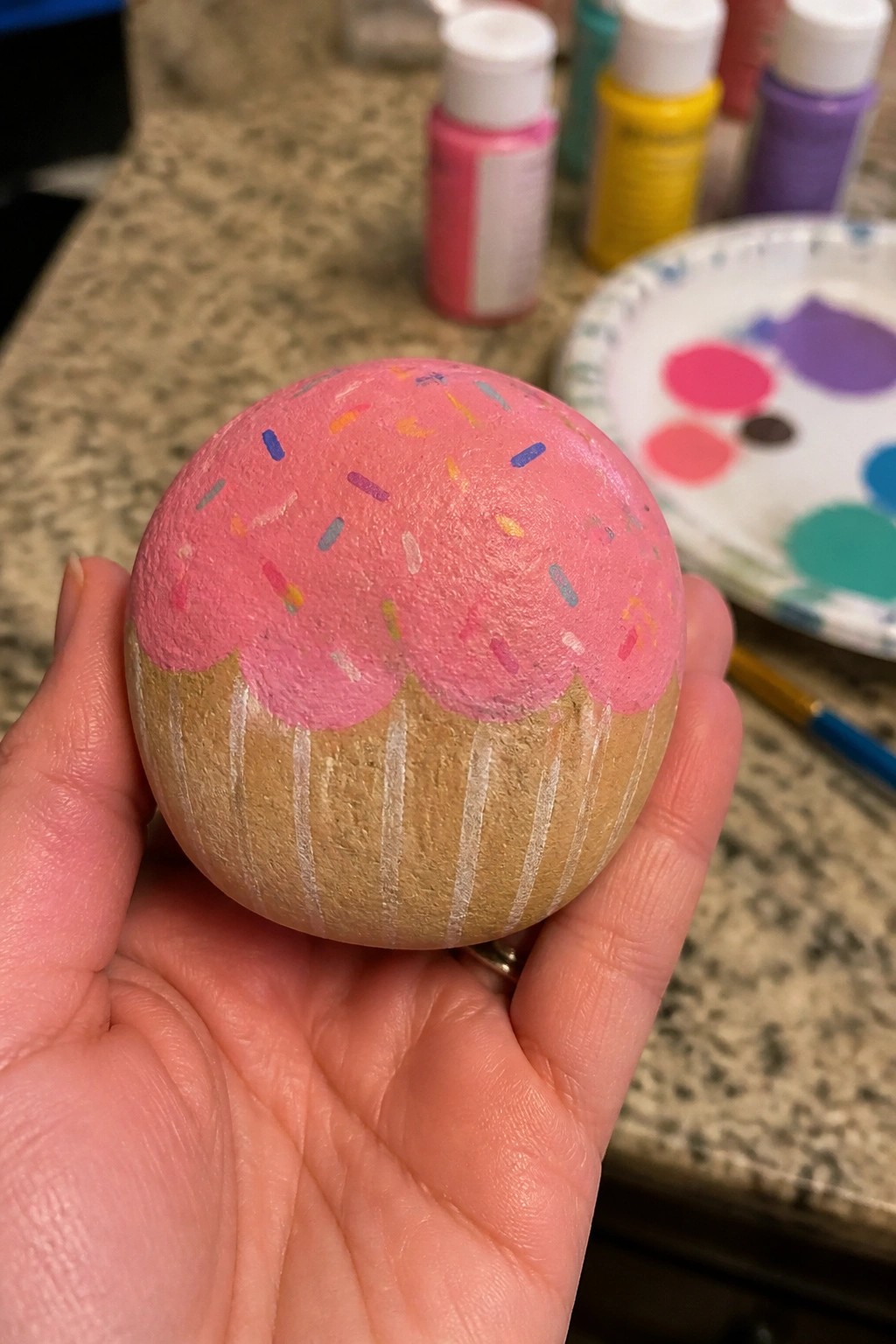 Hand holding a painted rock shaped like a cupcake with pink frosting and sprinkles.