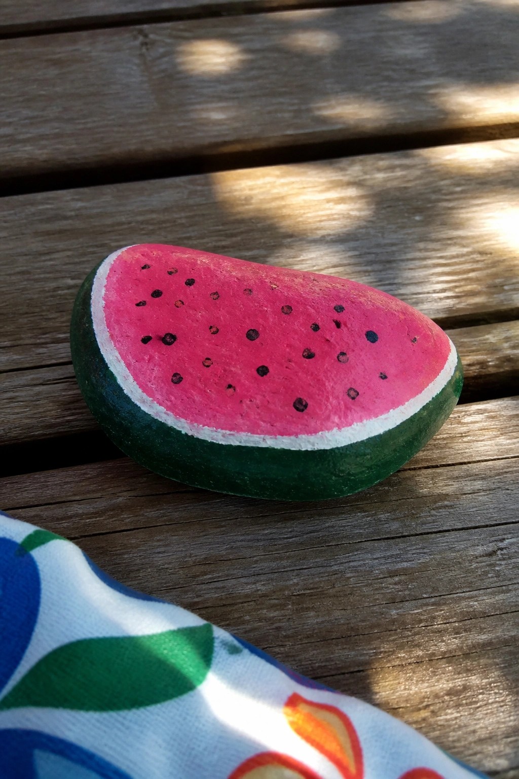 Watermelon slice painted on rock on sunlit wooden planks beside patterned fabric