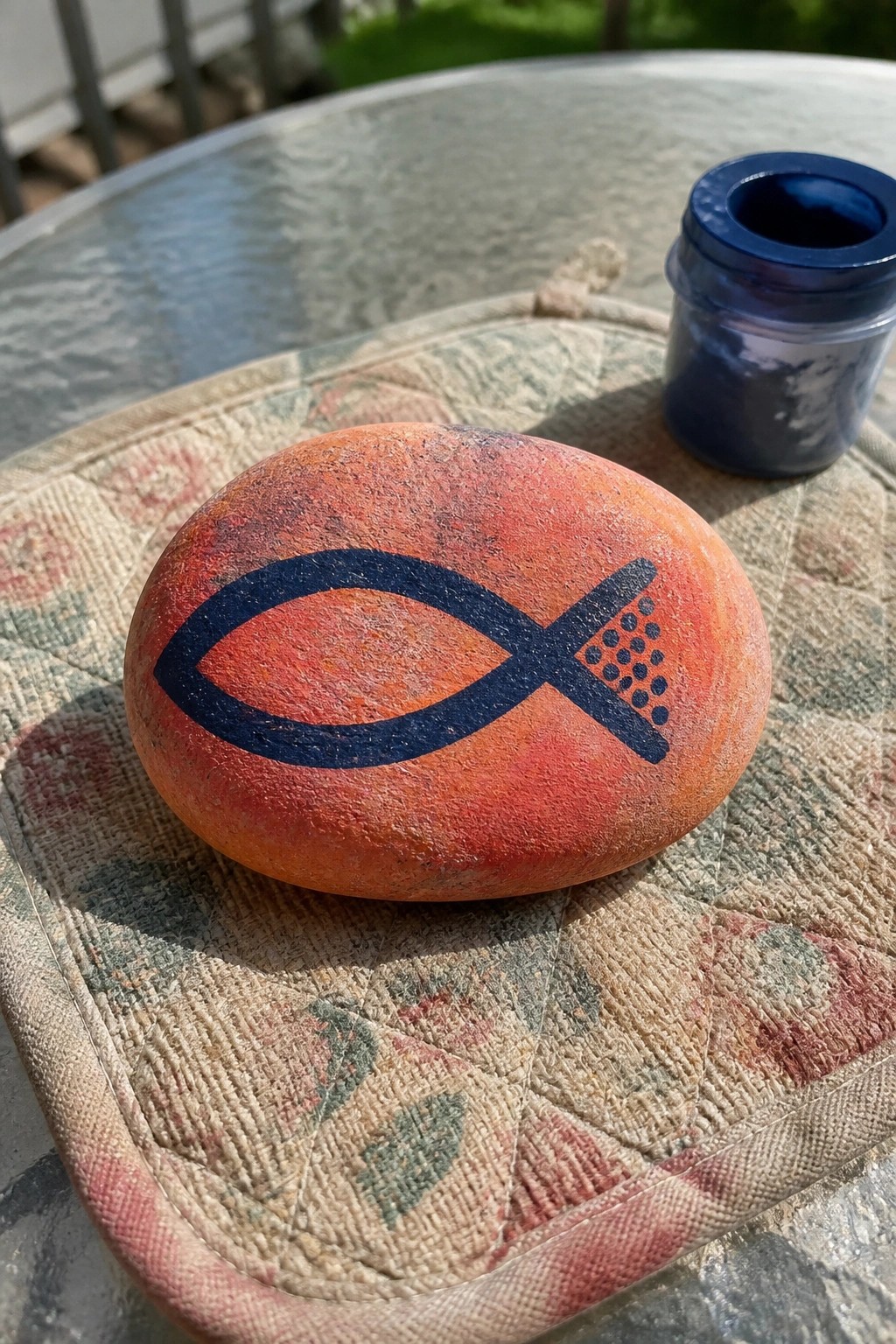 Orange rock with blue fish symbol on floral quilted mat beside blue jar