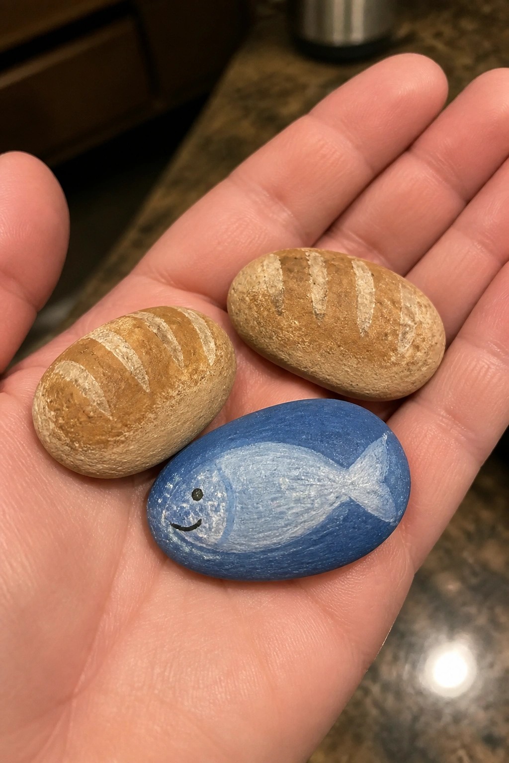 Hand holding two brown bread-shaped rocks and one smiling blue fish rock.