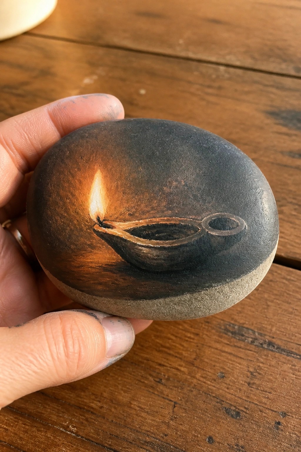 Hand holding smooth stone painted with glowing lit diya oil lamp