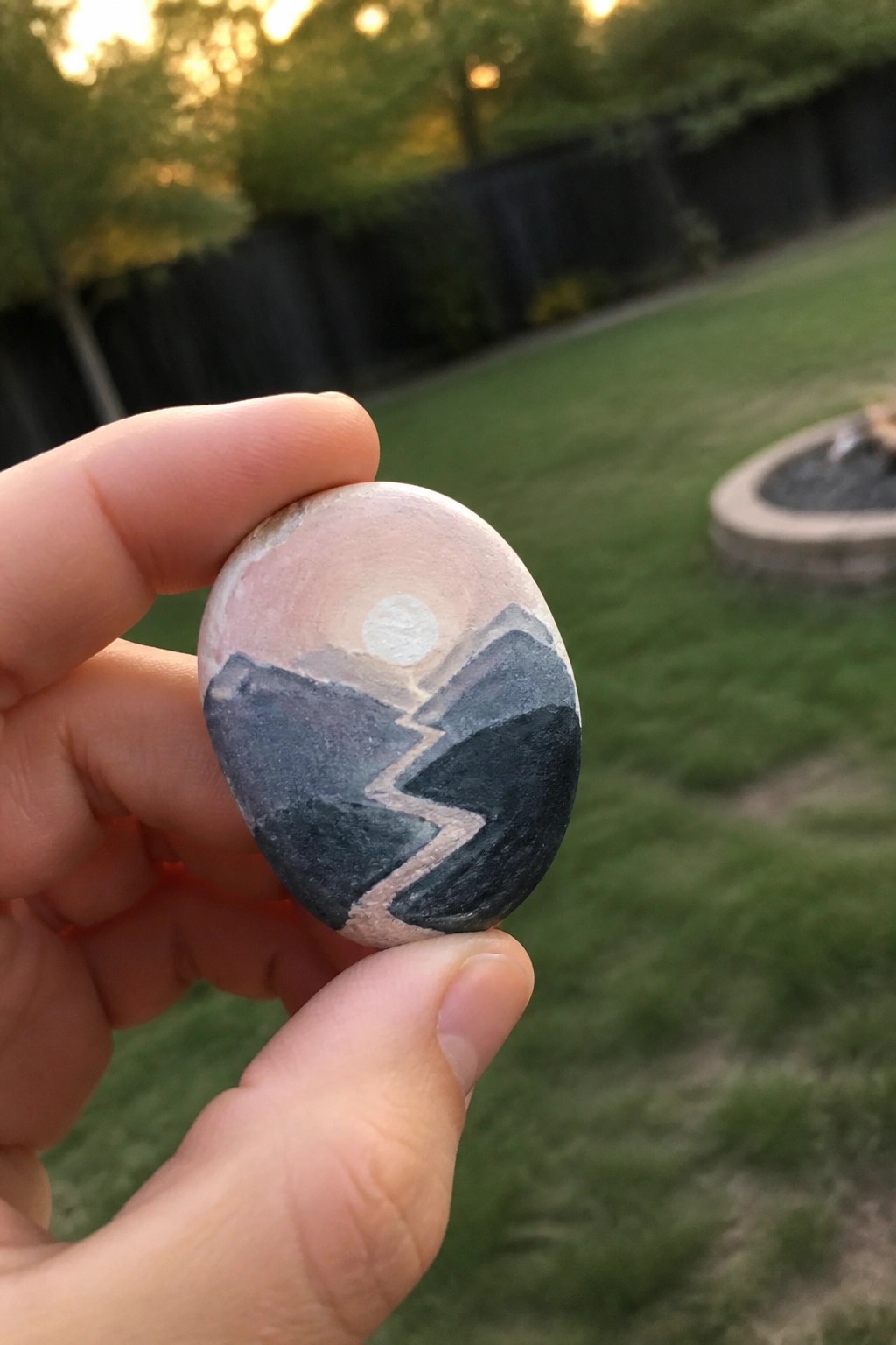 Hand holding painted stone with black mountains, white zigzag path, and pink sun.