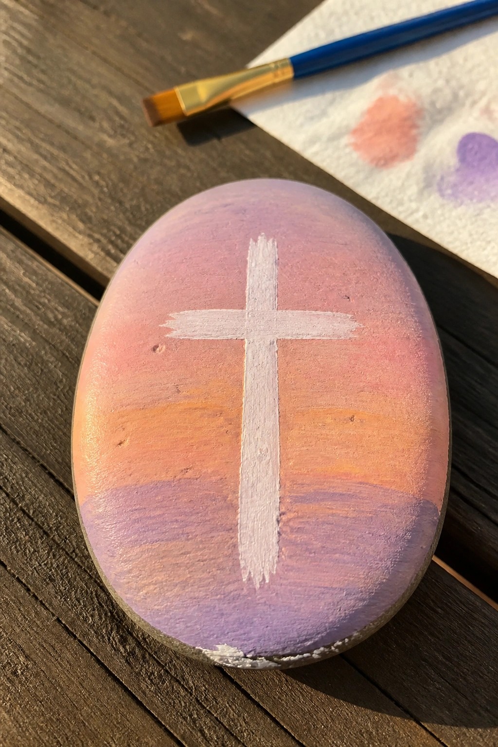 Painted rock with white cross over purple-pink-orange sunset gradient beside paintbrush.