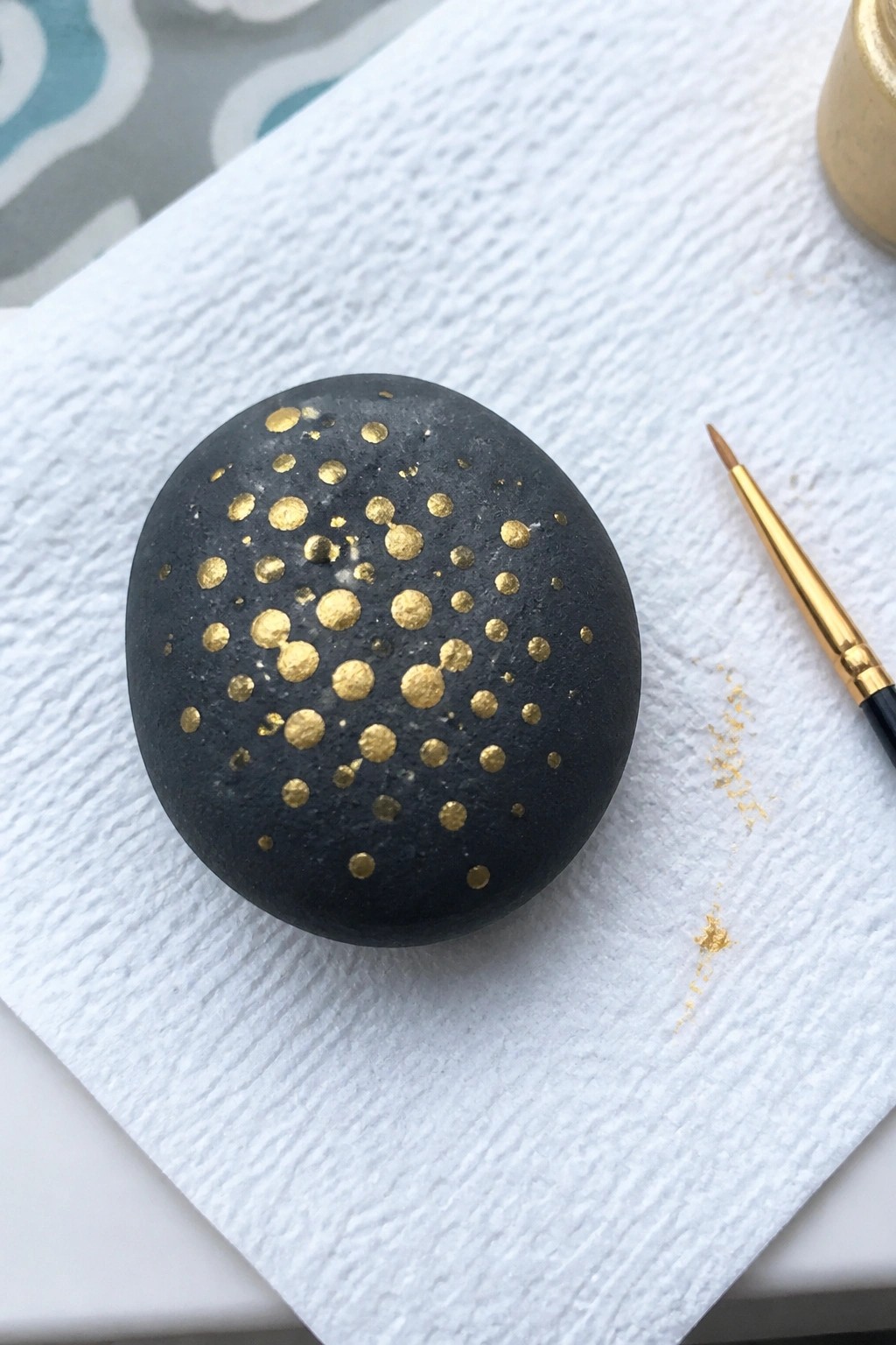 Black rock with gold dots on white paper towel beside fine paintbrush