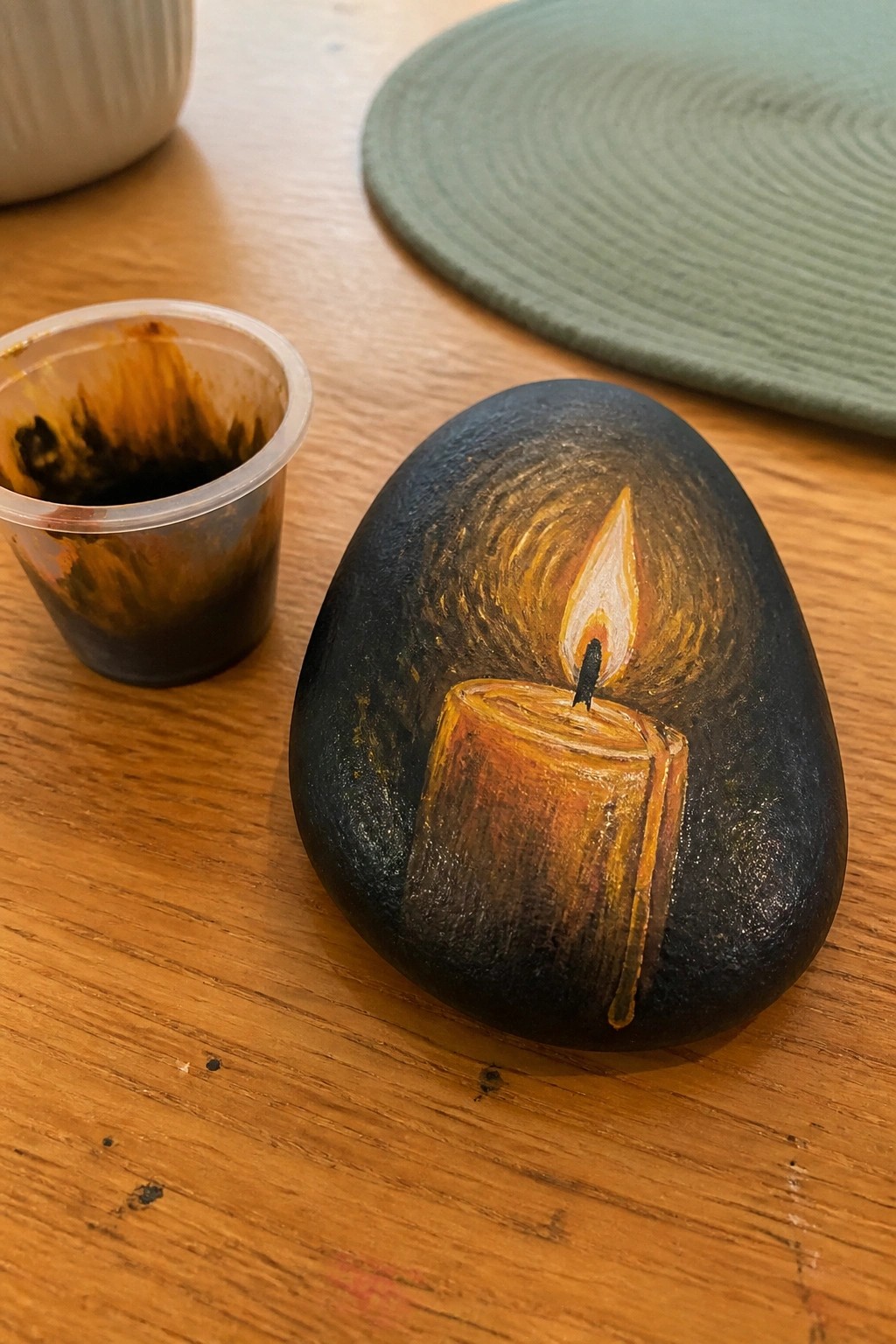 Black stone painted with glowing lit candle on wooden table beside paint cup