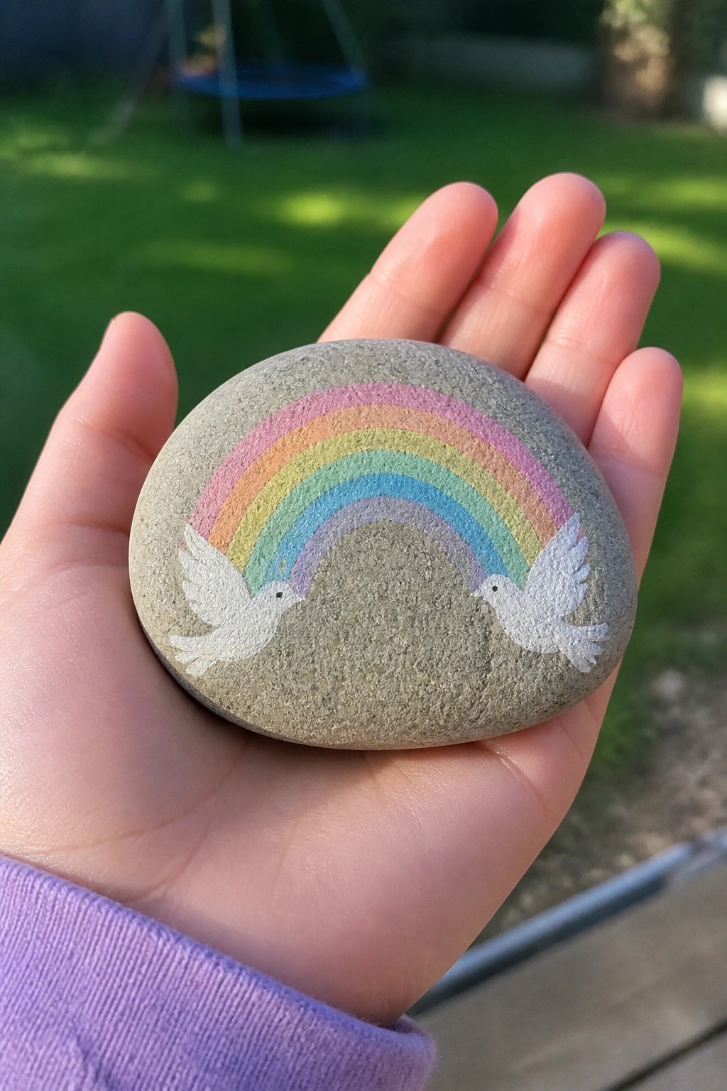 Hand in purple sleeve holding stone painted with rainbow and two white doves