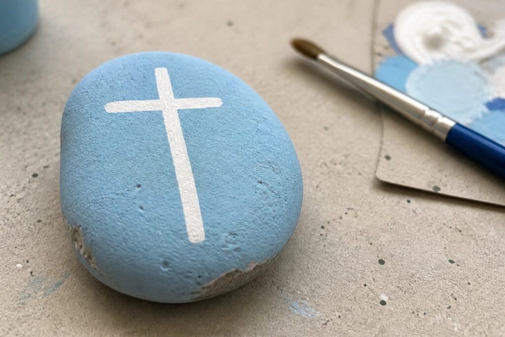 Light blue painted rock with white cross beside paintbrush on splattered surface.
