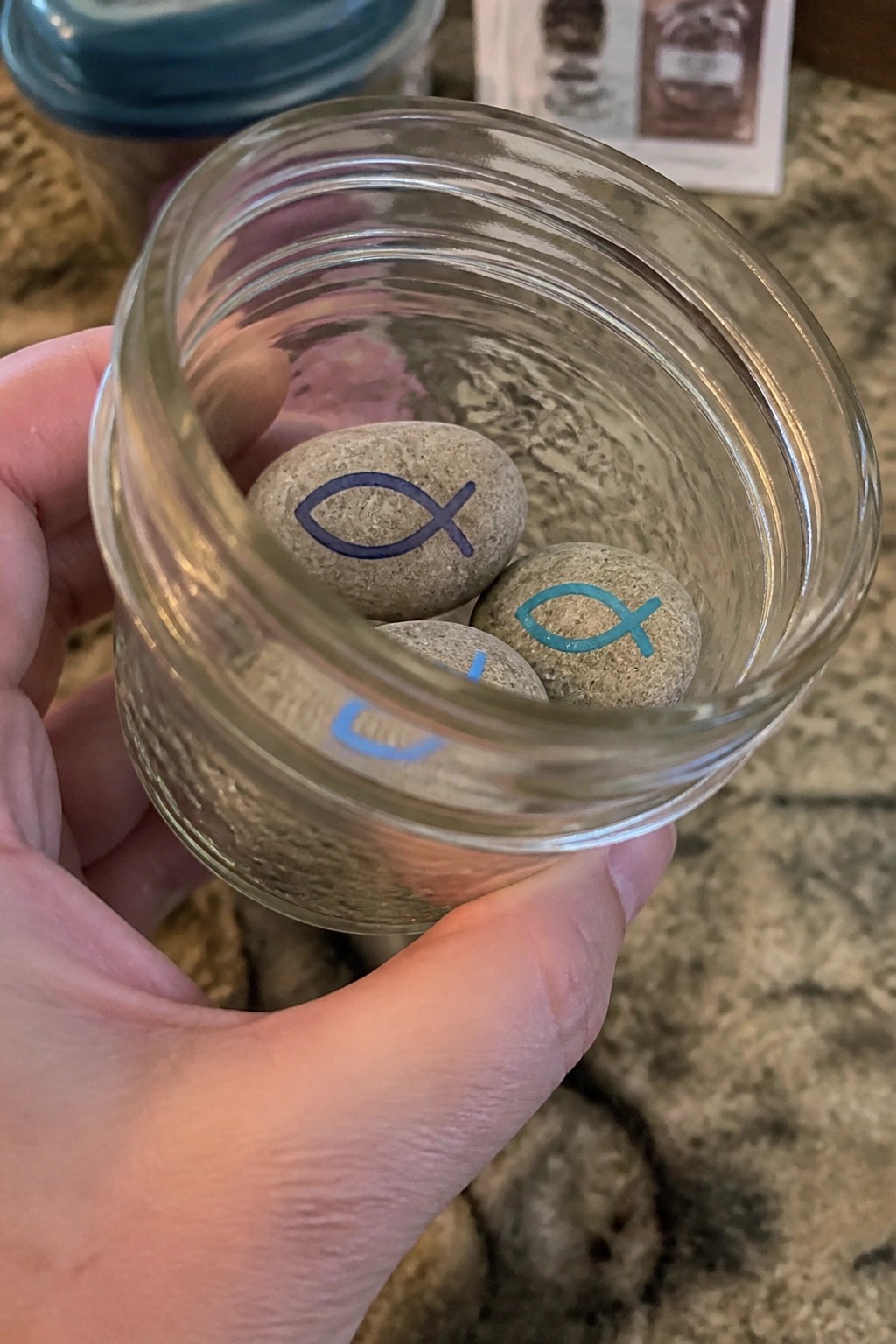Hand holding open glass jar with three stones painted with colorful fish symbols