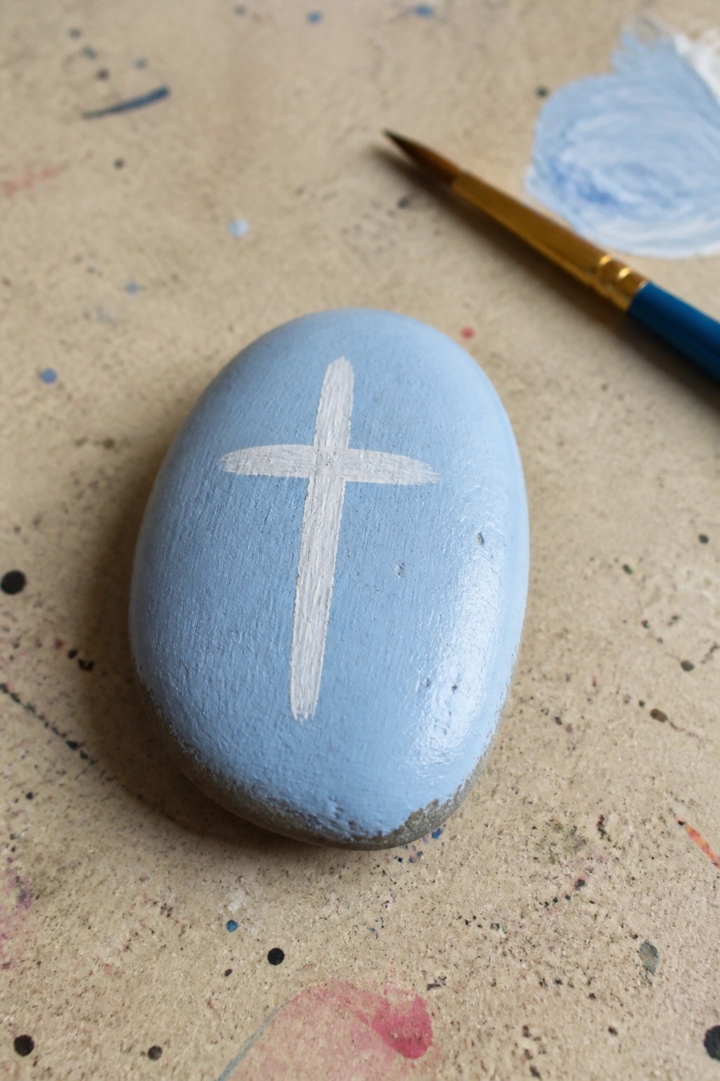 Light blue painted rock with white cross beside paintbrush on splattered surface.