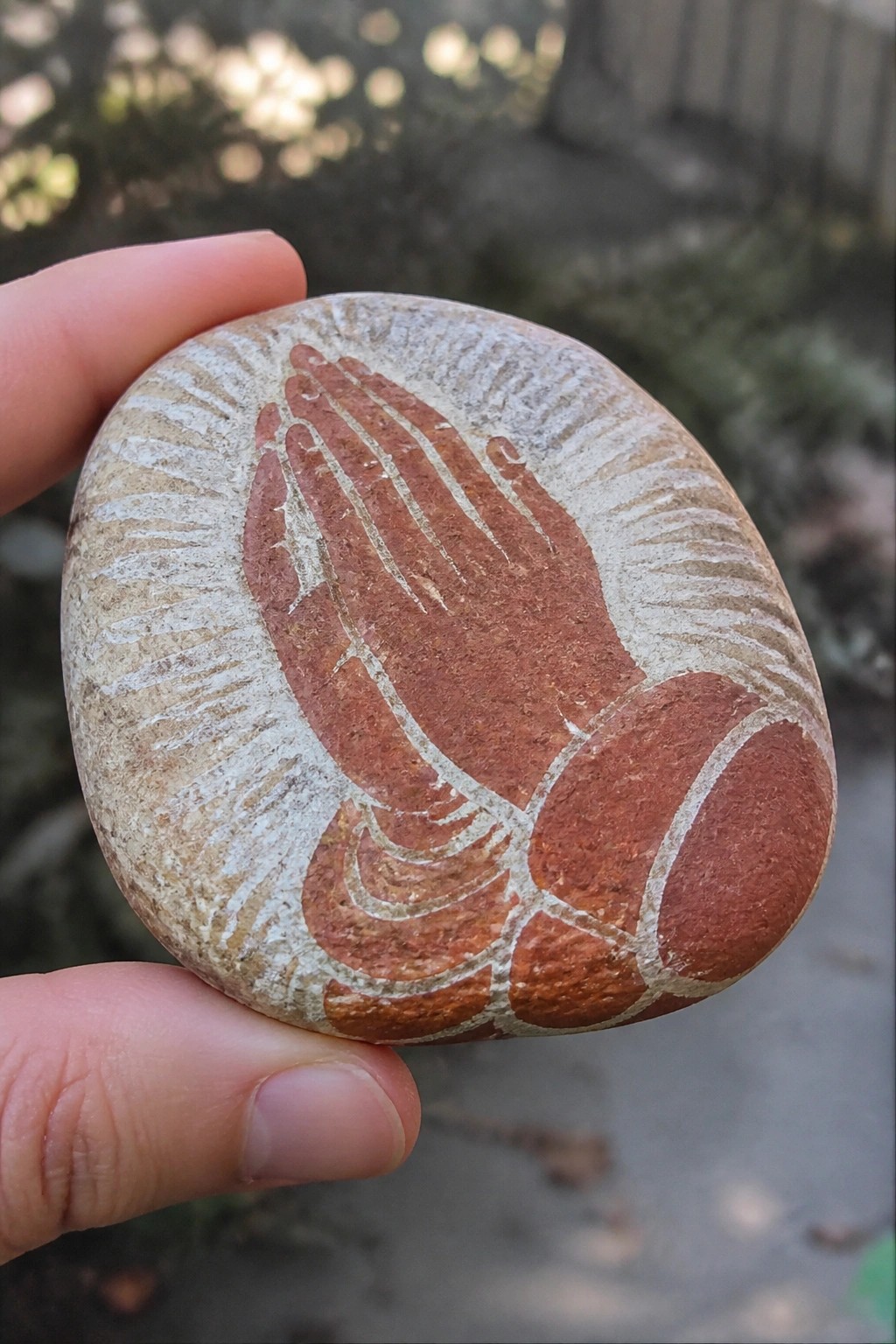 Hand holding oval stone painted with reddish praying hands and white rays