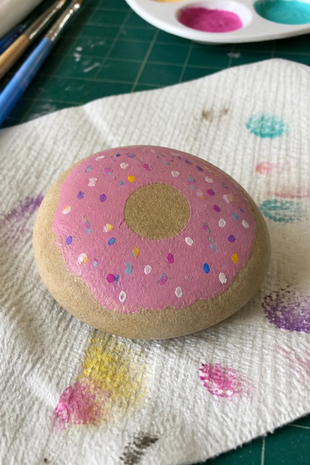 A smooth oval stone painted with pink icing and colorful sprinkles to resemble a donut sits on a paper towel.