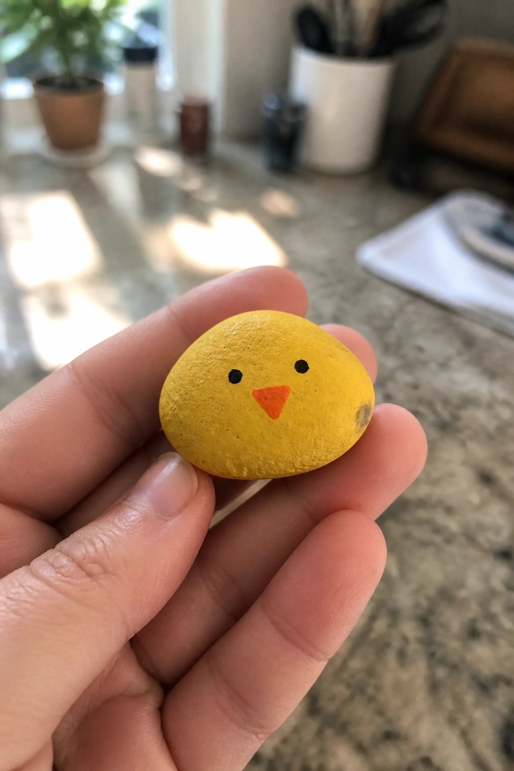 Hand holding small yellow painted rock with black eyes and orange chick beak