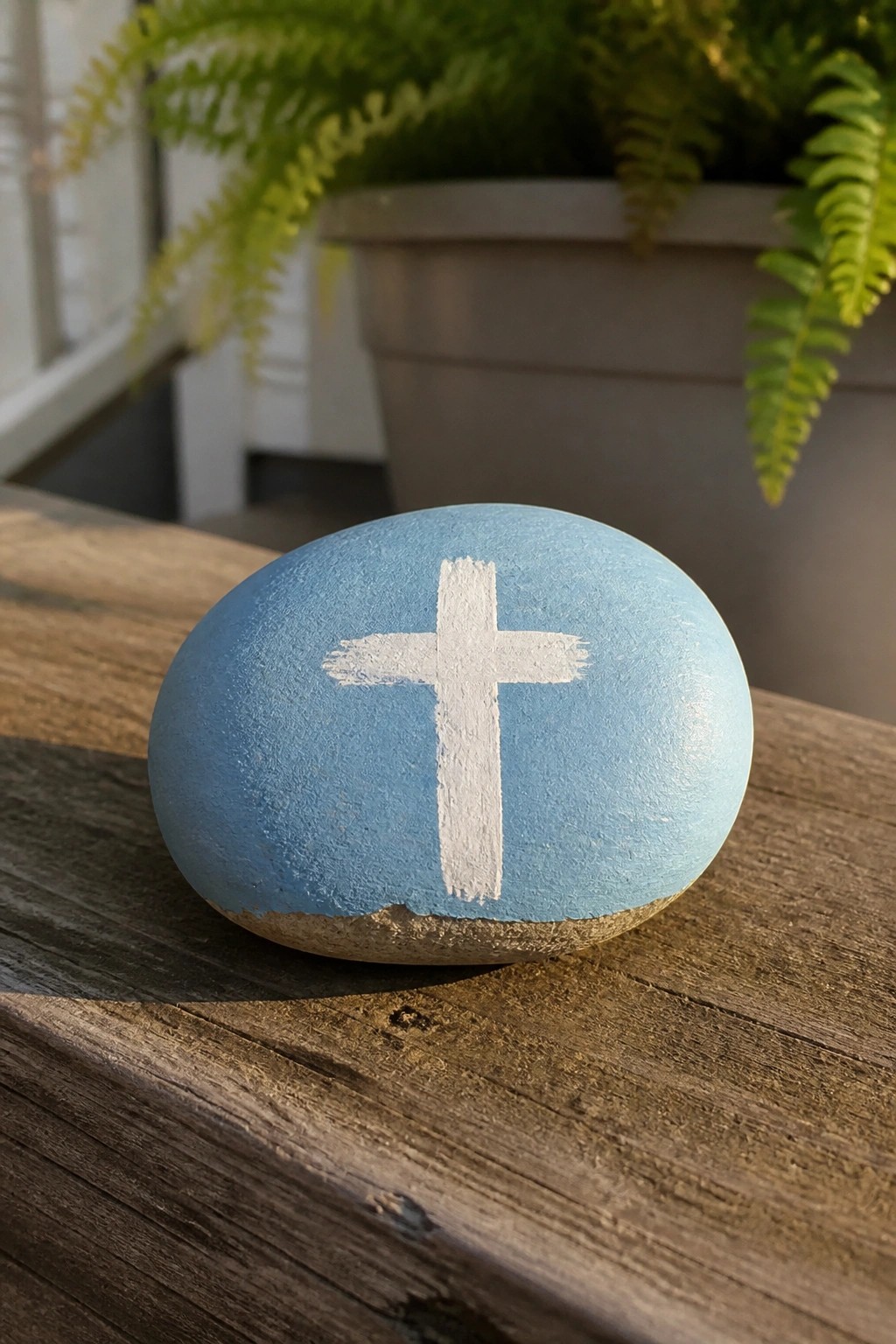 Light blue painted rock with white cross on wooden table near plant pot