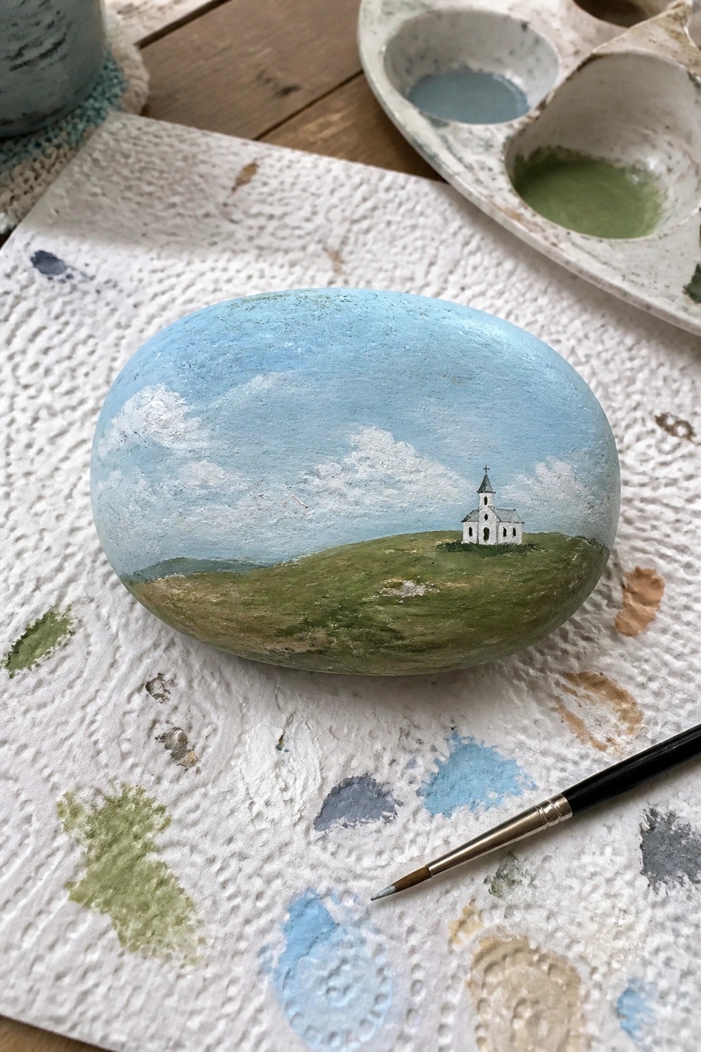 Painted rock showing white church on green hills under blue cloudy sky with supplies nearby