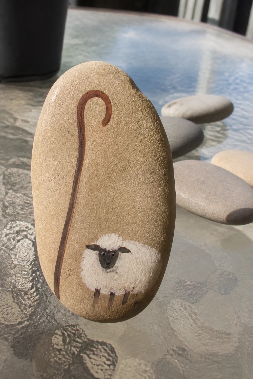 Beige oval stone painted with white sheep, black face, and brown crook.