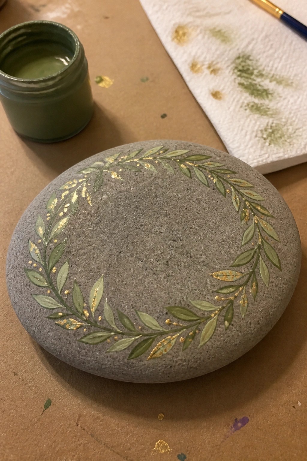 Gray stone with green and gold leafy wreath beside paint jar on craft table