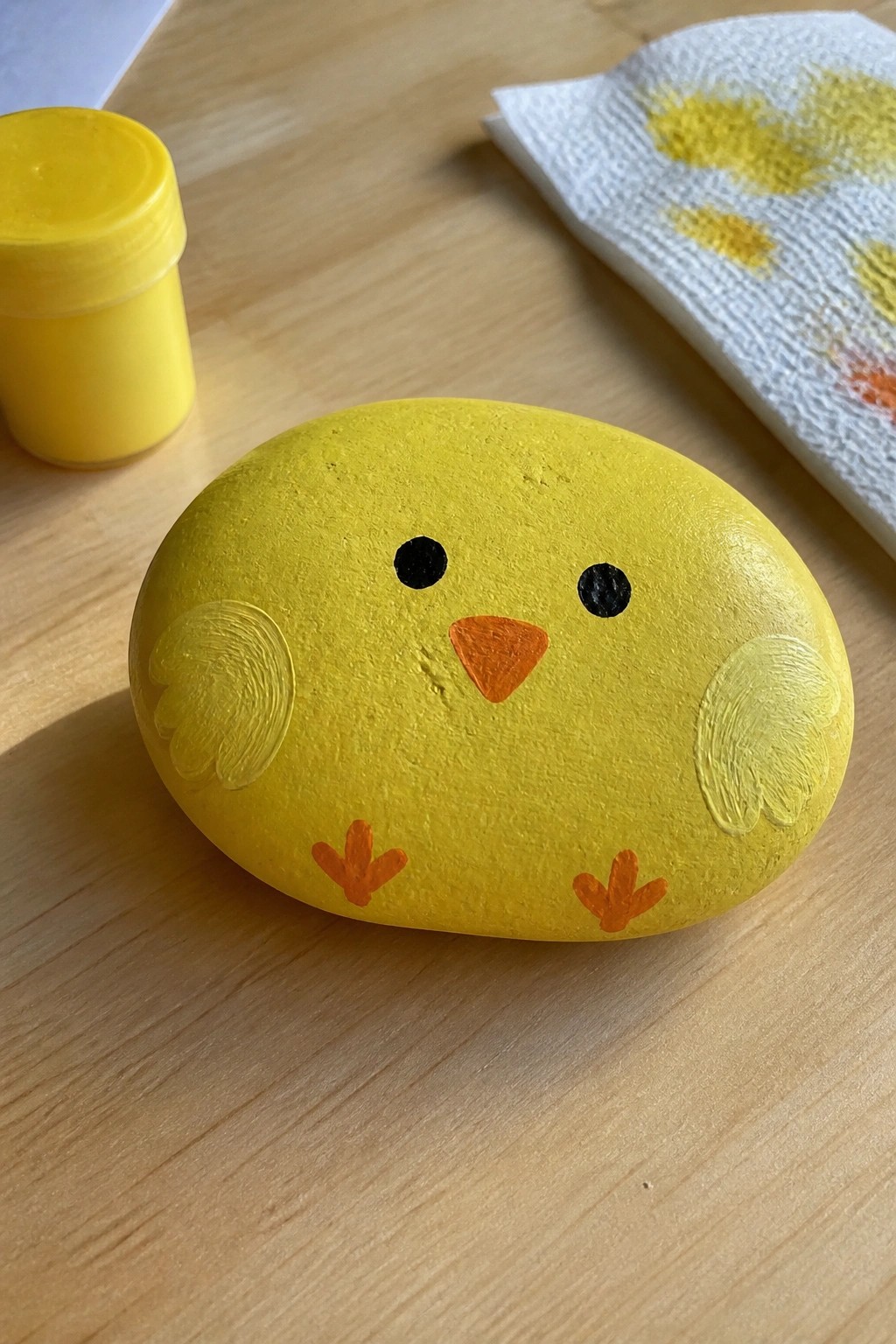 Yellow painted chick rock with orange beak, feet, and black eyes beside paint jar
