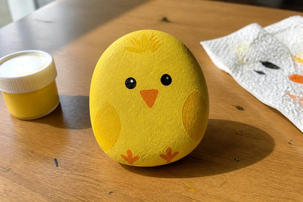 24 Fun Rock Painting Ideas for Kids and Family Craft Days Yellow painted chick rock with orange beak, feet, and black eyes beside paint jar