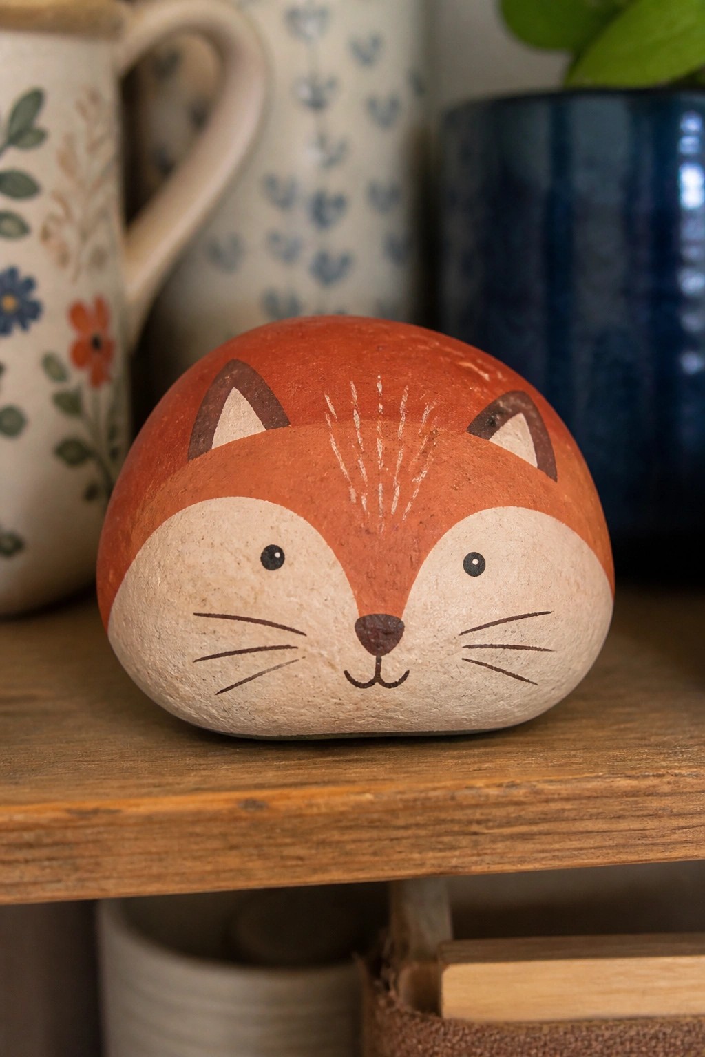 Painted fox on orange-beige stone atop wooden shelf with pottery behind