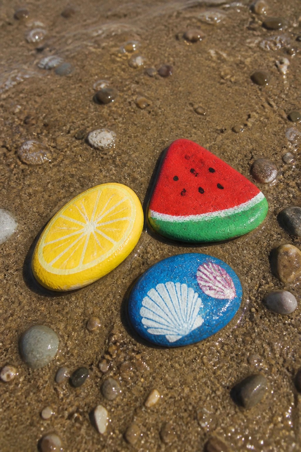 Three painted rocks on wet sand: lemon, watermelon slice, and seashells.