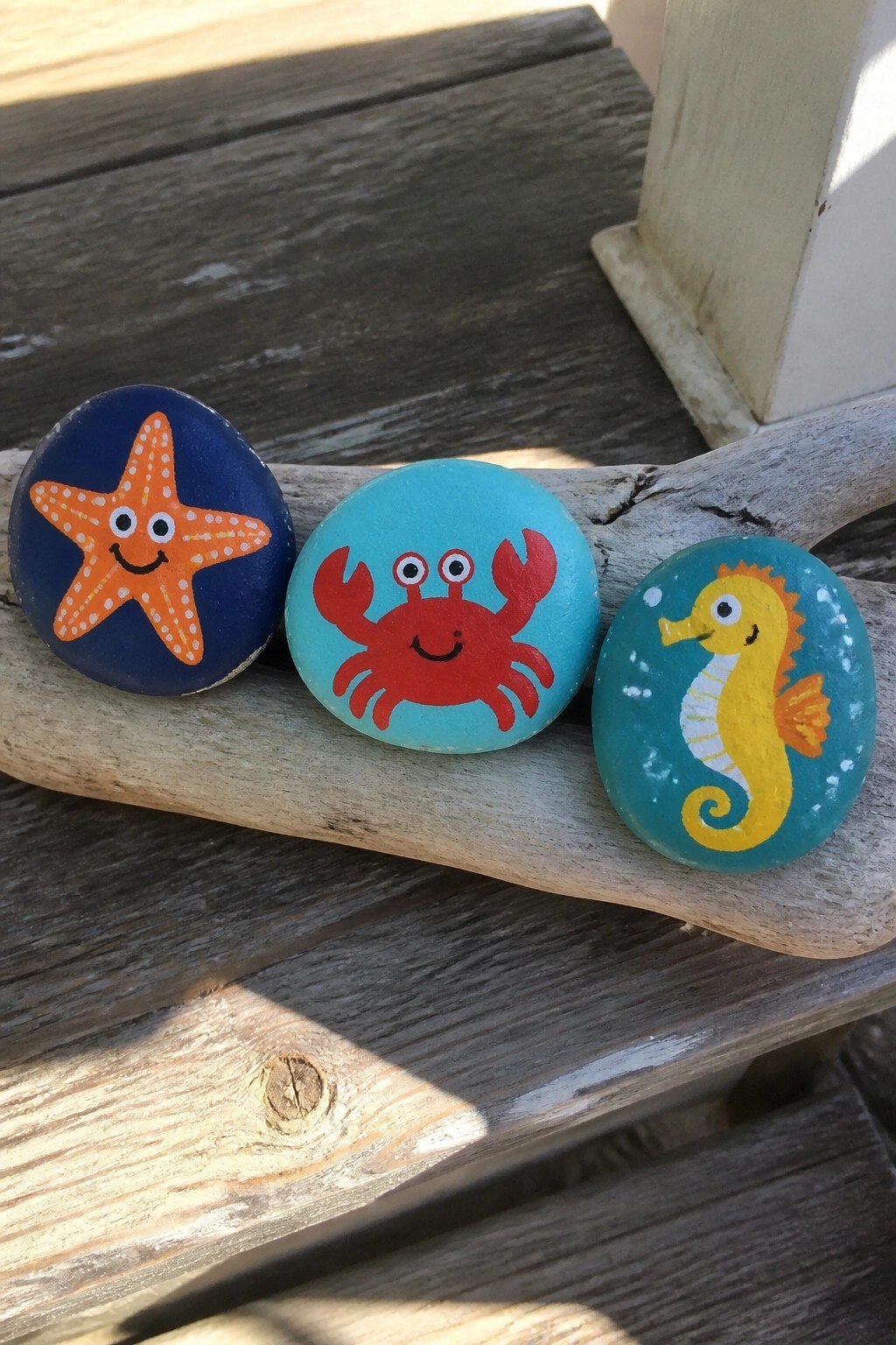Three painted rocks with smiling starfish, crab, and seahorse on weathered wood.