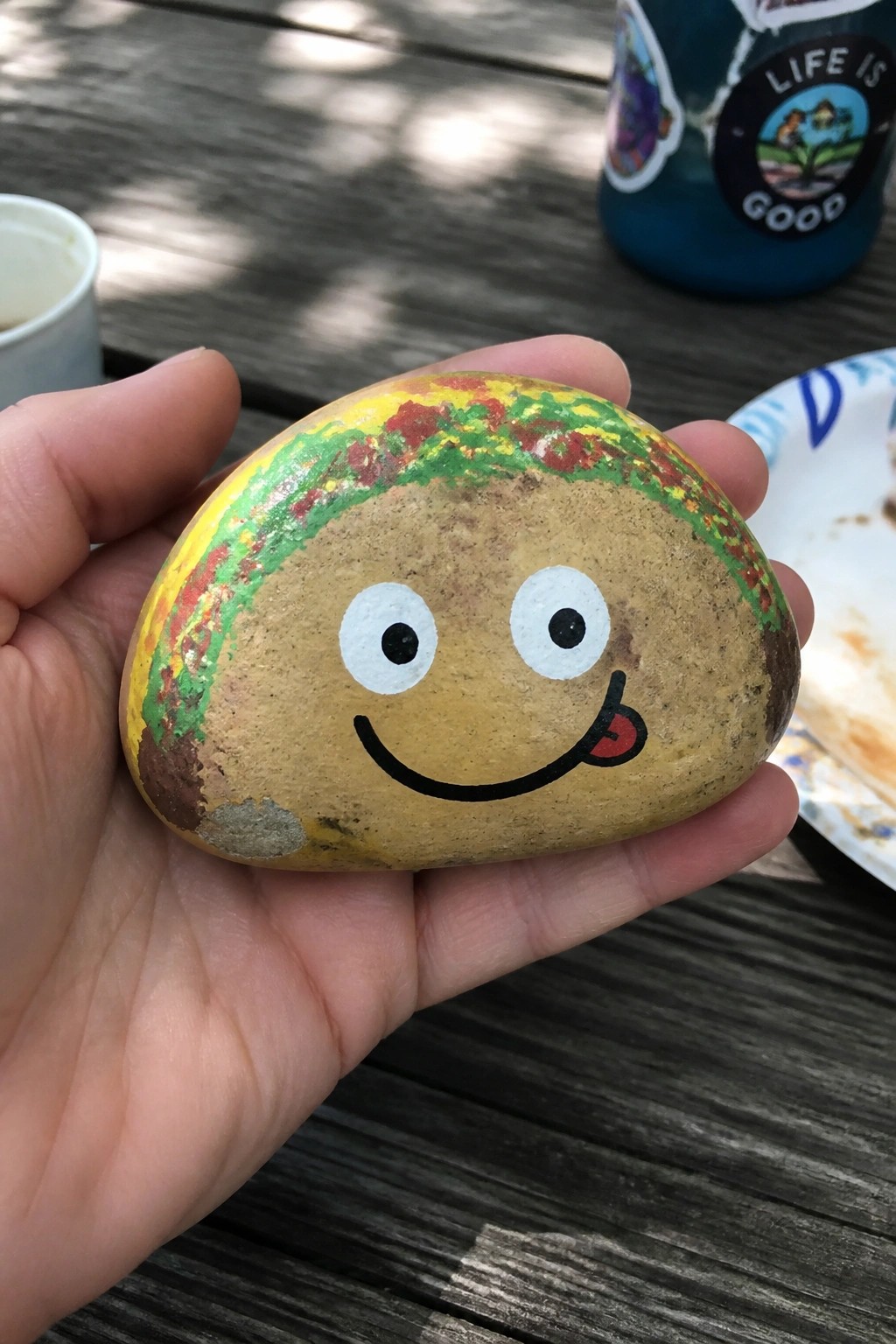 Hand holding smiling painted rock with white eyes and red tongue on wooden table.