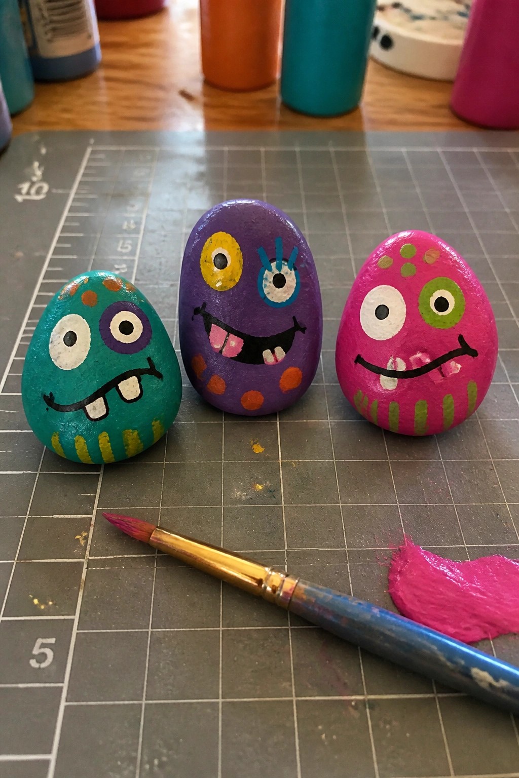 Three colorful monster-faced rocks on a gridded mat beside a paintbrush and pink paint.