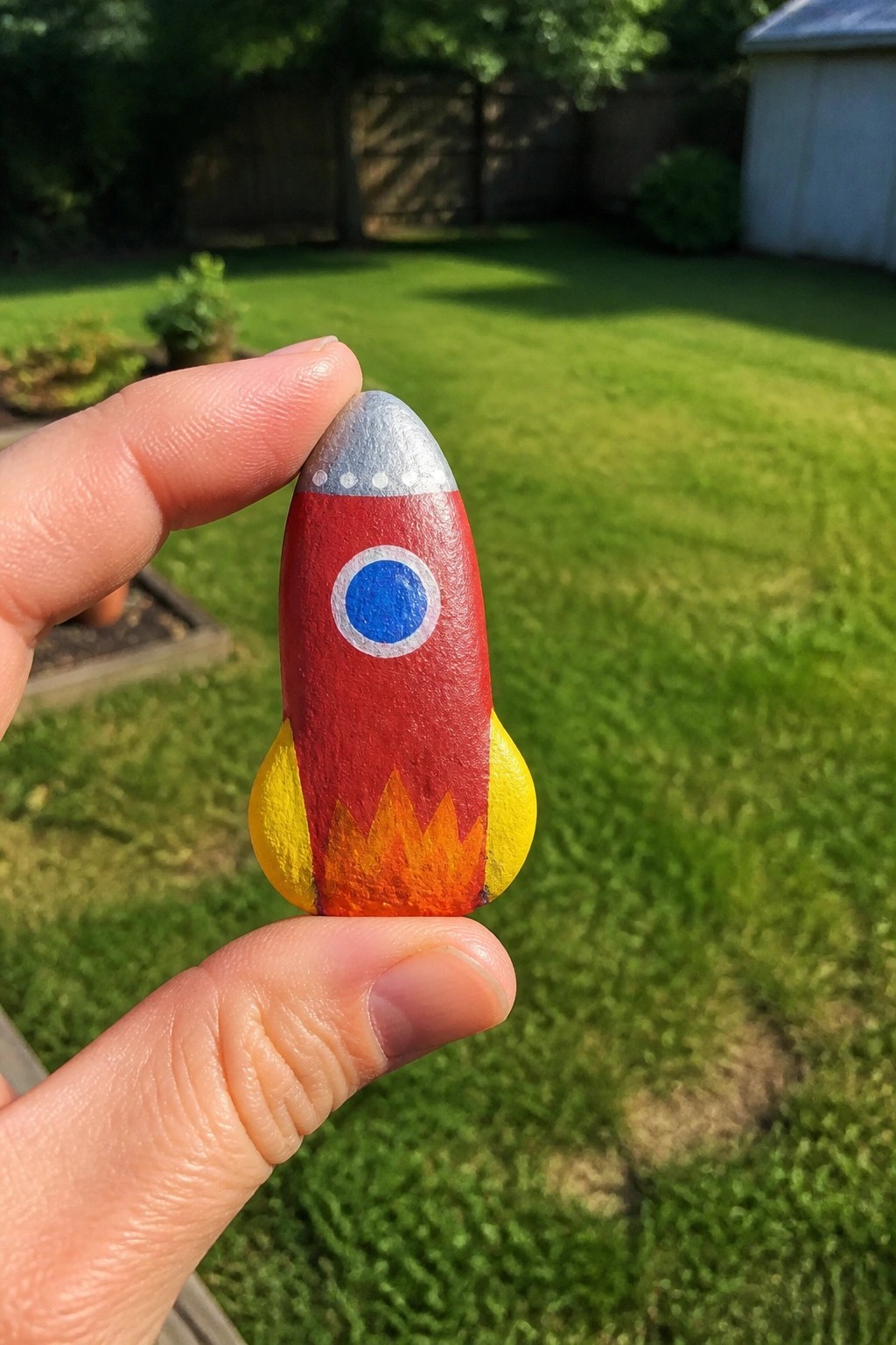 A hand holds a small painted rock shaped and colored like a red rocket with a silver top, blue window, yellow fins, and flame details against a grassy backyard.