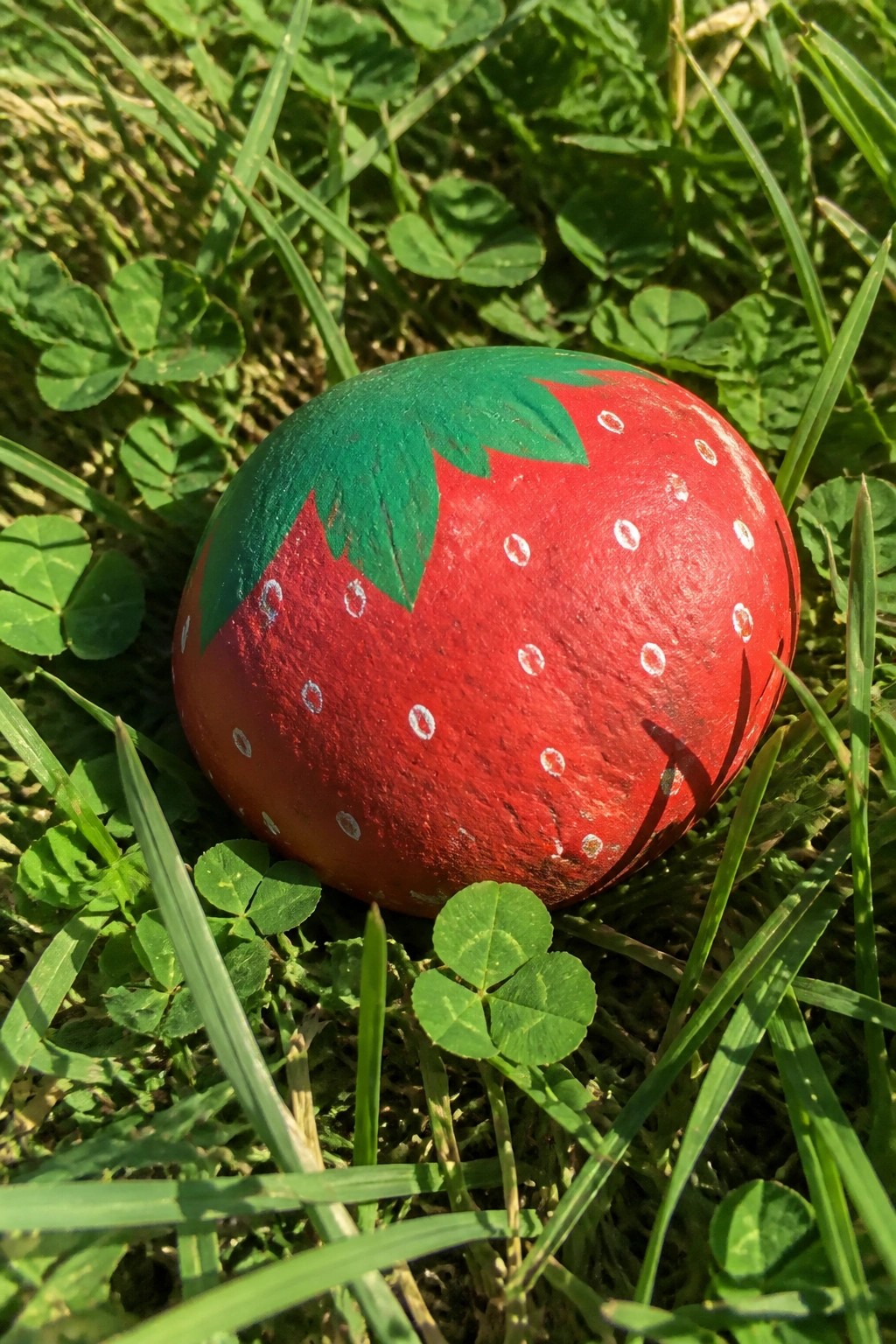 Painted red strawberry rock with green leaves and seeds in grass