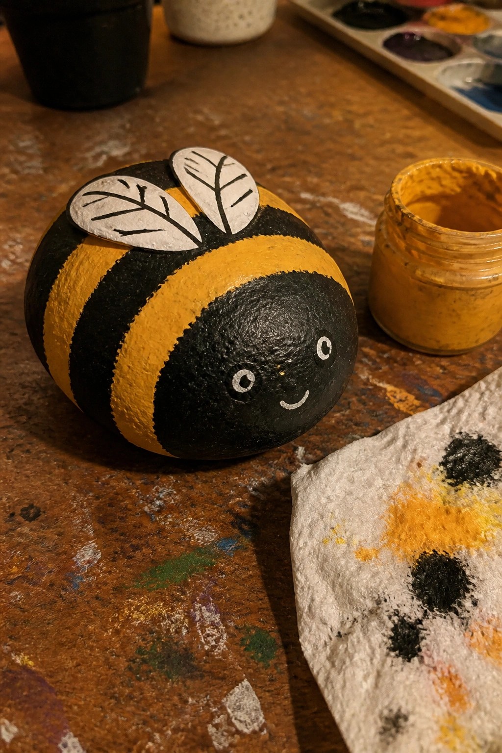 Painted bumblebee rock with yellow-black stripes and wings beside paint jar on wood