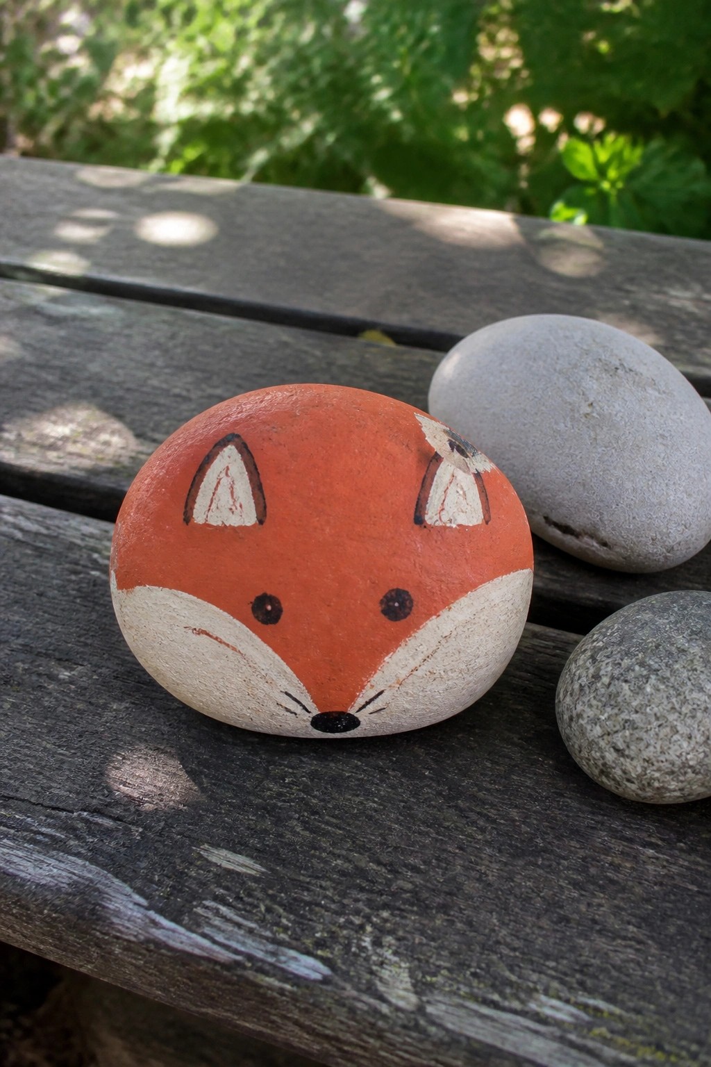 A rounded orange and white painted rock shaped like a fox face sits on a wooden surface next to two plain gray stones.