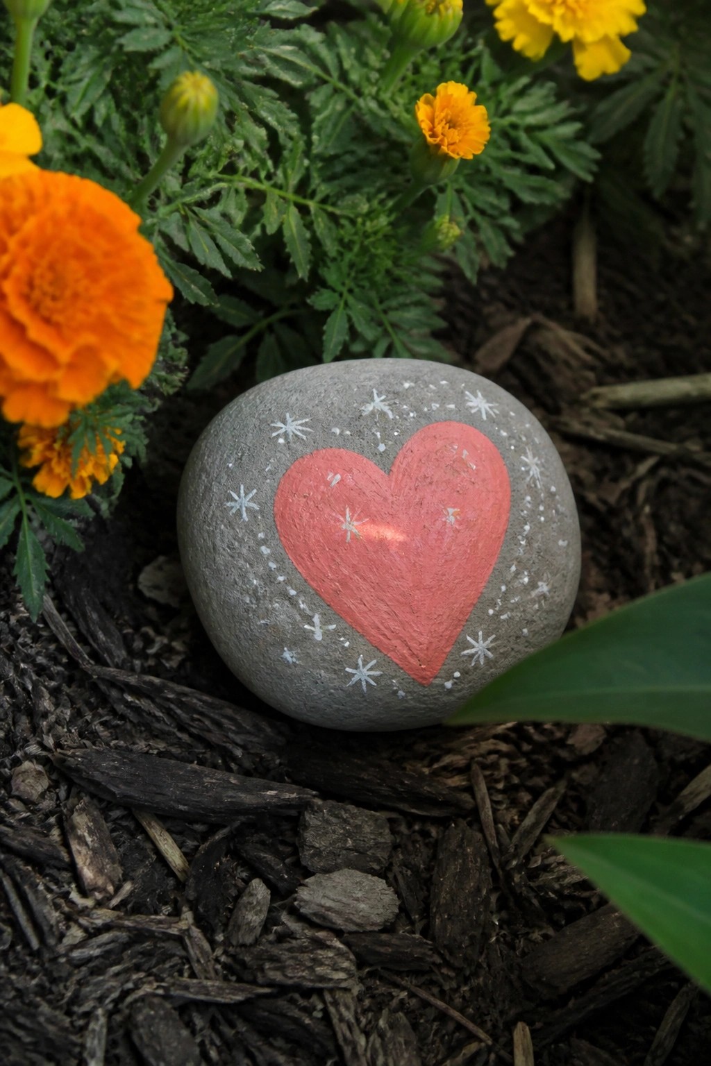 Gray rock painted with red heart and white stars amid orange marigolds.