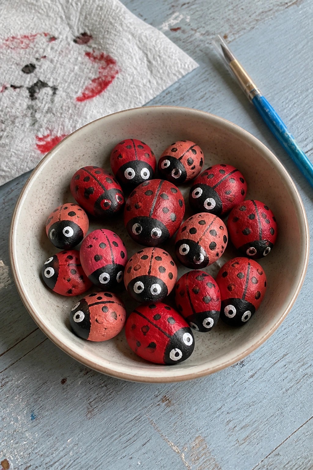 Bowl of red and black painted ladybug rocks beside paintbrush on blue wood