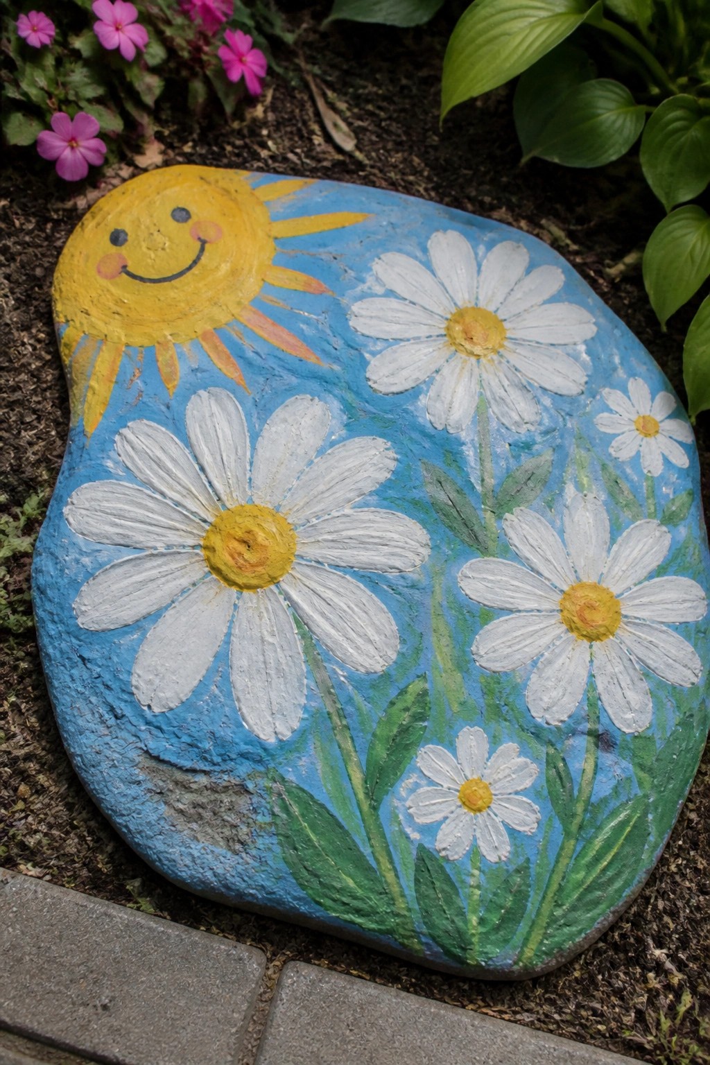 Painted rock with smiling yellow sun, white daisies, and blue background.