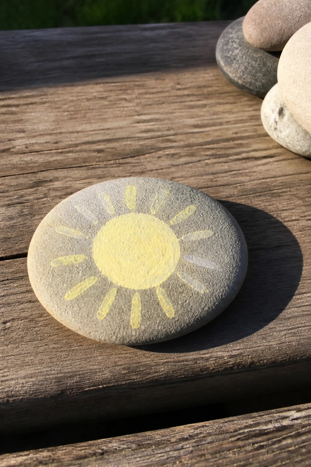 Gray stone with yellow sun painted on it rests on wooden surface near other pebbles