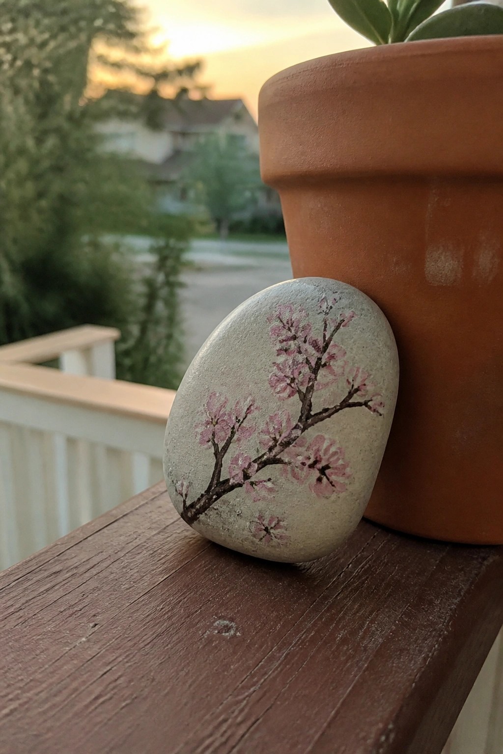 Painted rock with pink cherry blossoms beside terracotta pot on wooden railing at sunset