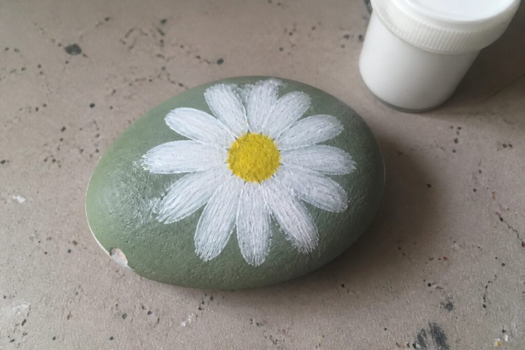Gray-blue painted rock with white daisy flower and yellow center beside paint jar