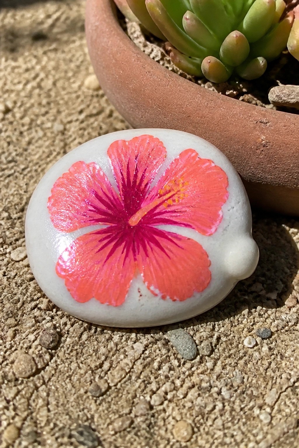 White stone painted with glossy pink hibiscus flower on sandy ground near terracotta pot.
