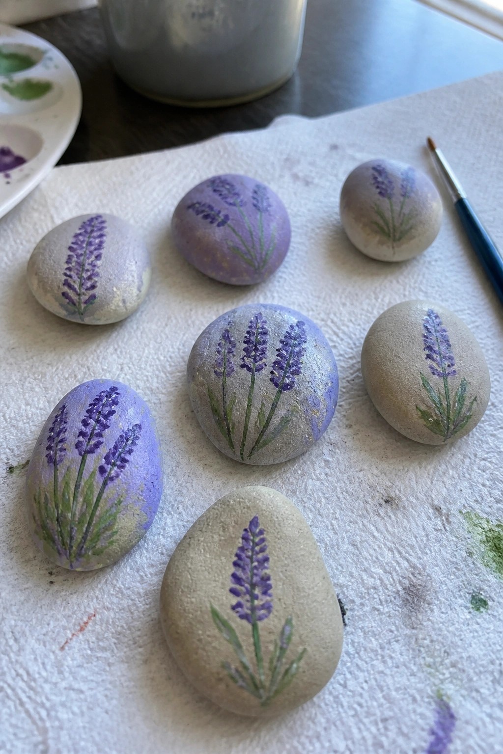 Lavender-painted rocks in purple and green on white paper towel with supplies nearby