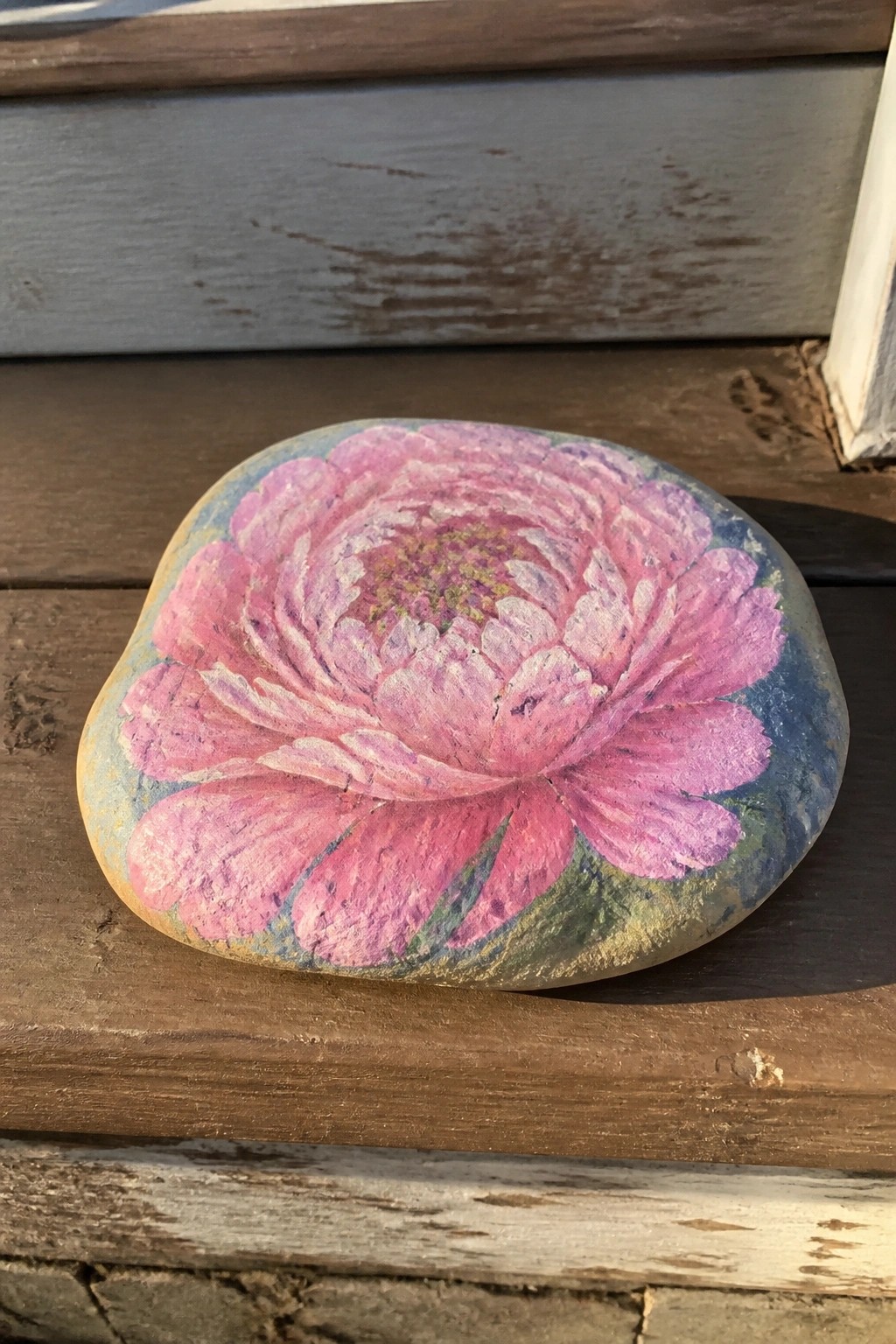 Hand-painted rock with large pink peony flower on wooden surface.