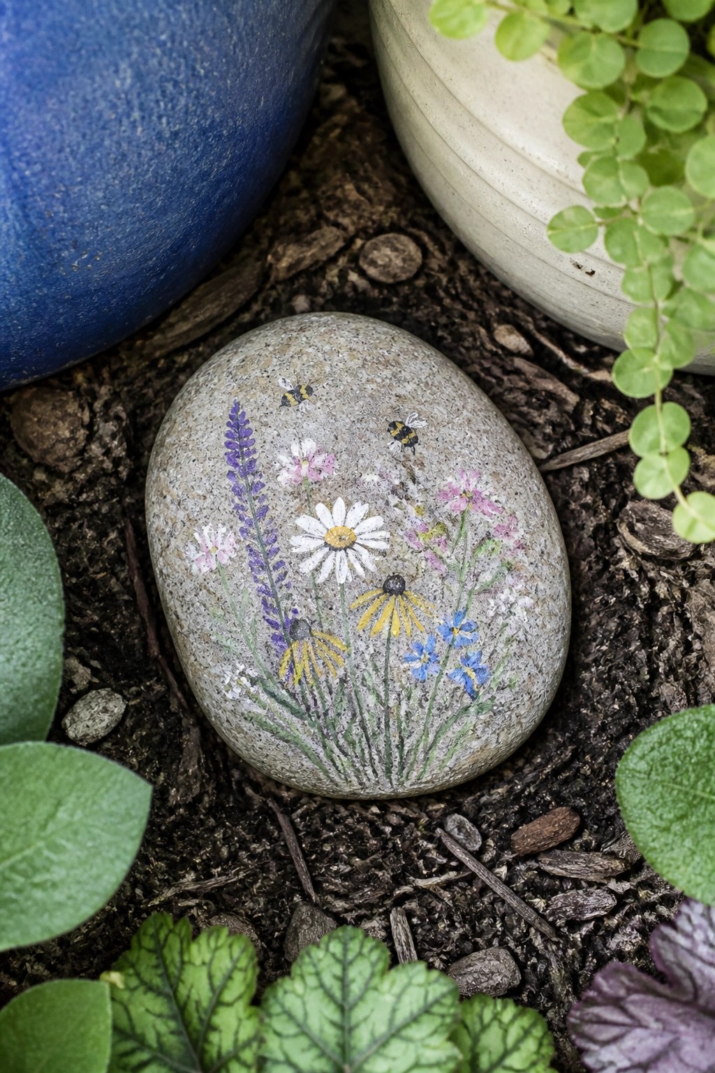 Painted rock with daisies, lavender, yellow flowers, blue blooms and two bees on soil