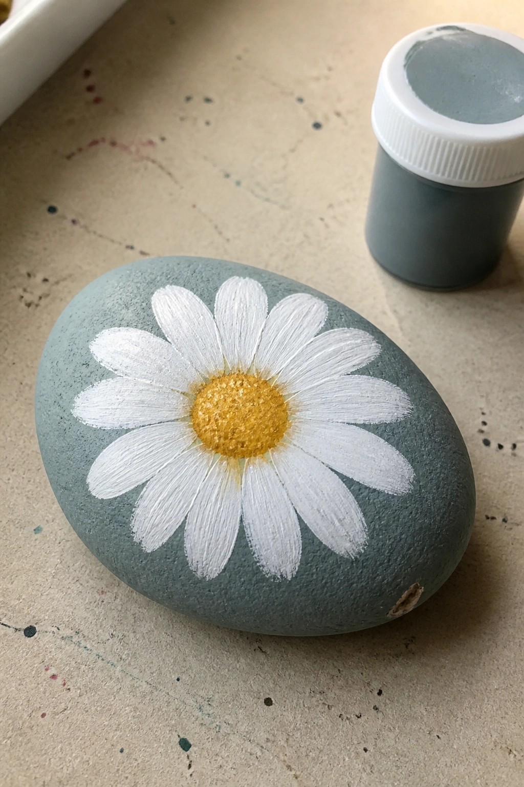 Gray-blue painted rock with white daisy flower and yellow center beside paint jar