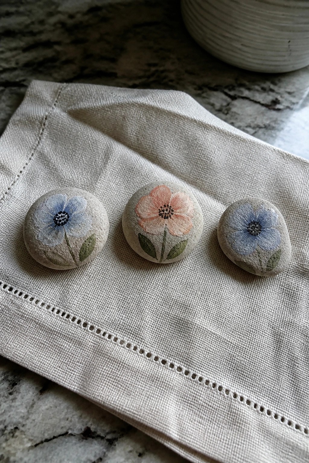 Three painted stones with blue and pink flowers on linen cloth