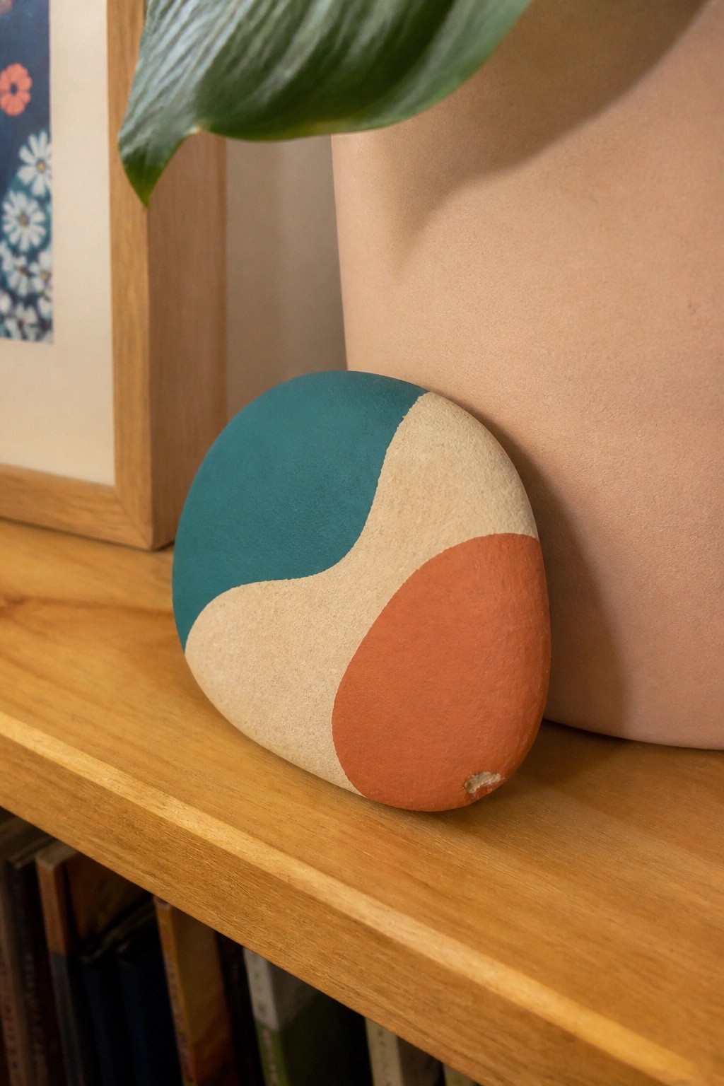 Abstract painted stone in teal, beige, and orange on wooden shelf by vase.