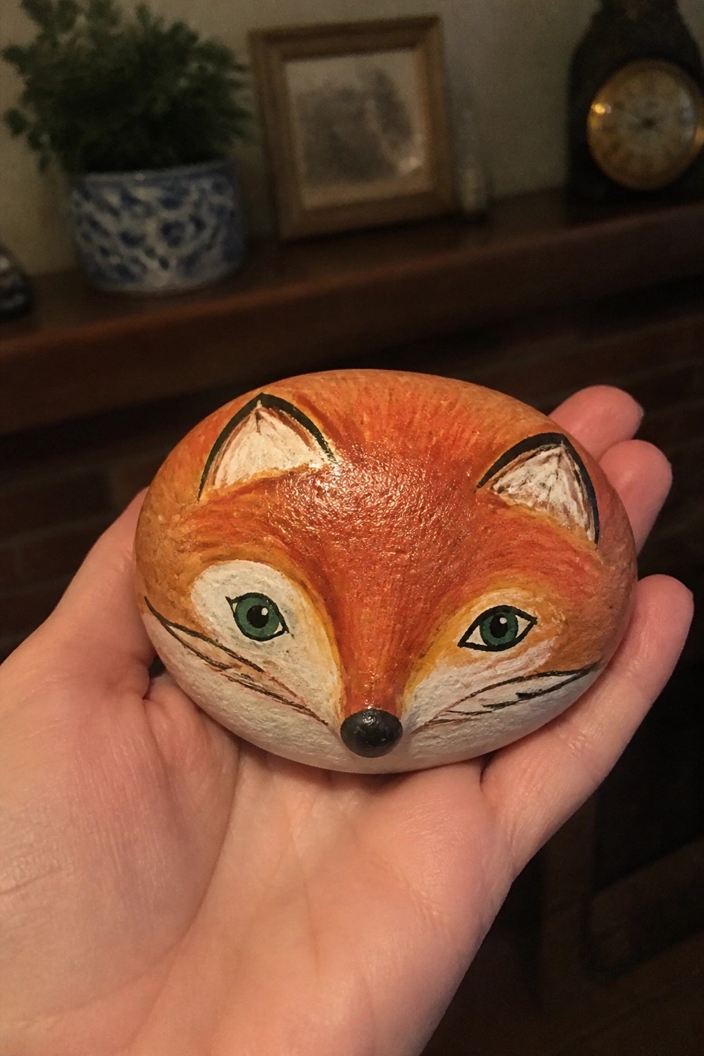 Hand holding orange fox-painted rock with green eyes, white muzzle, and black nose.
