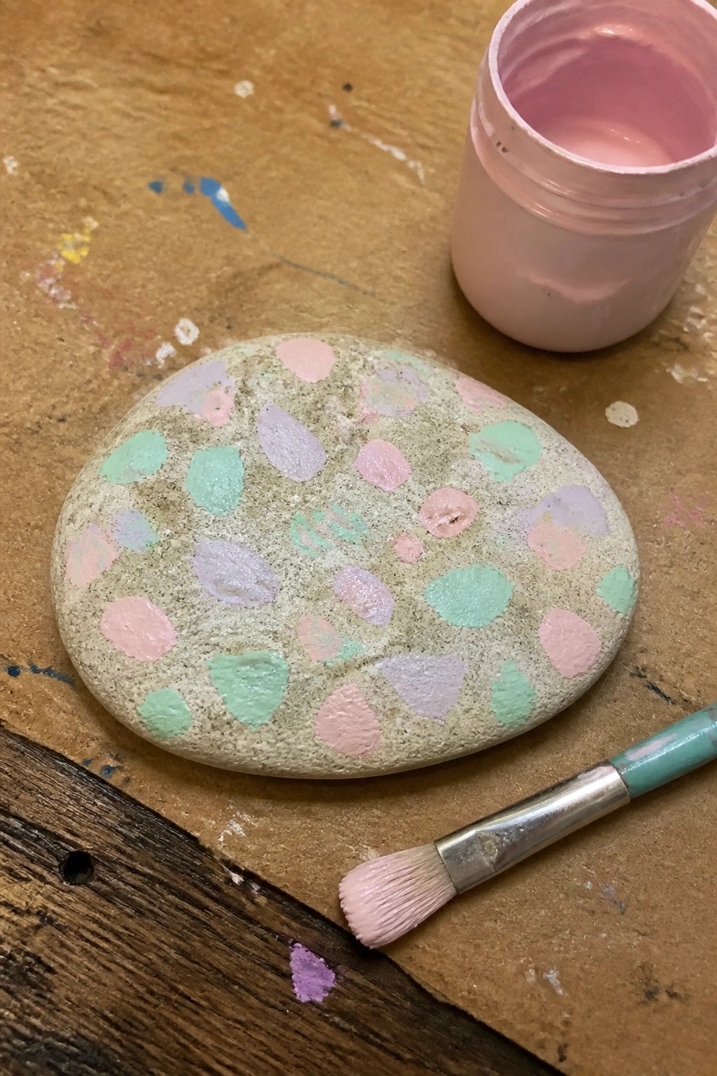 Painted stone with pastel dots beside pink paint jar and brush on wood