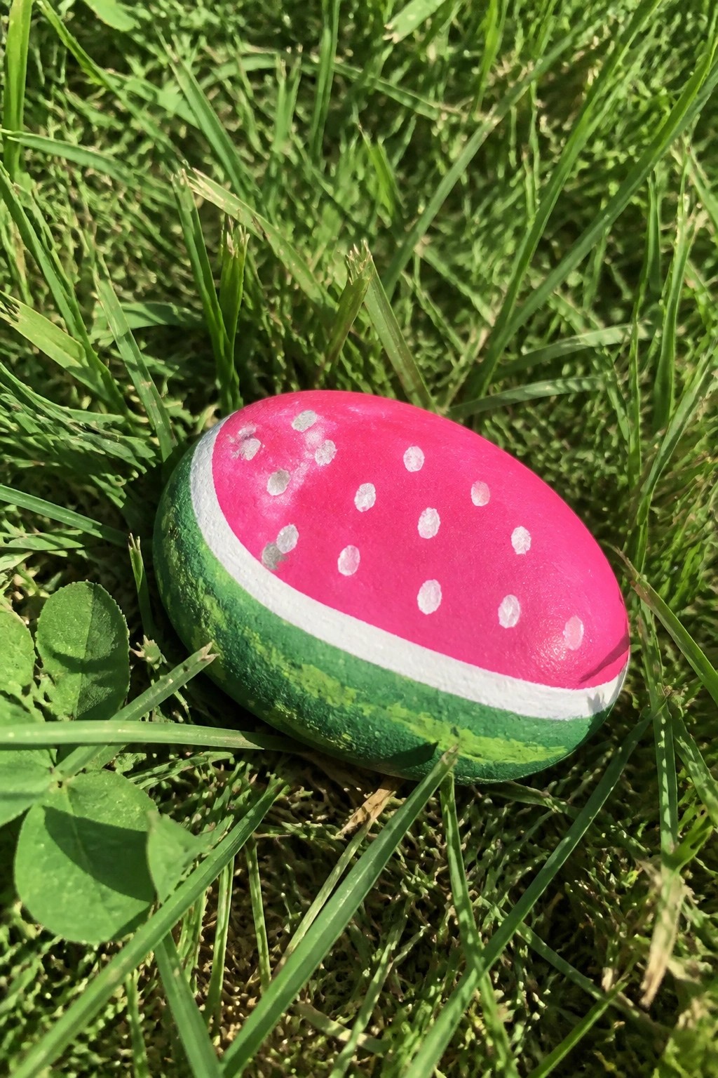 Watermelon-painted rock with pink top, white seeds, and green rind on grass