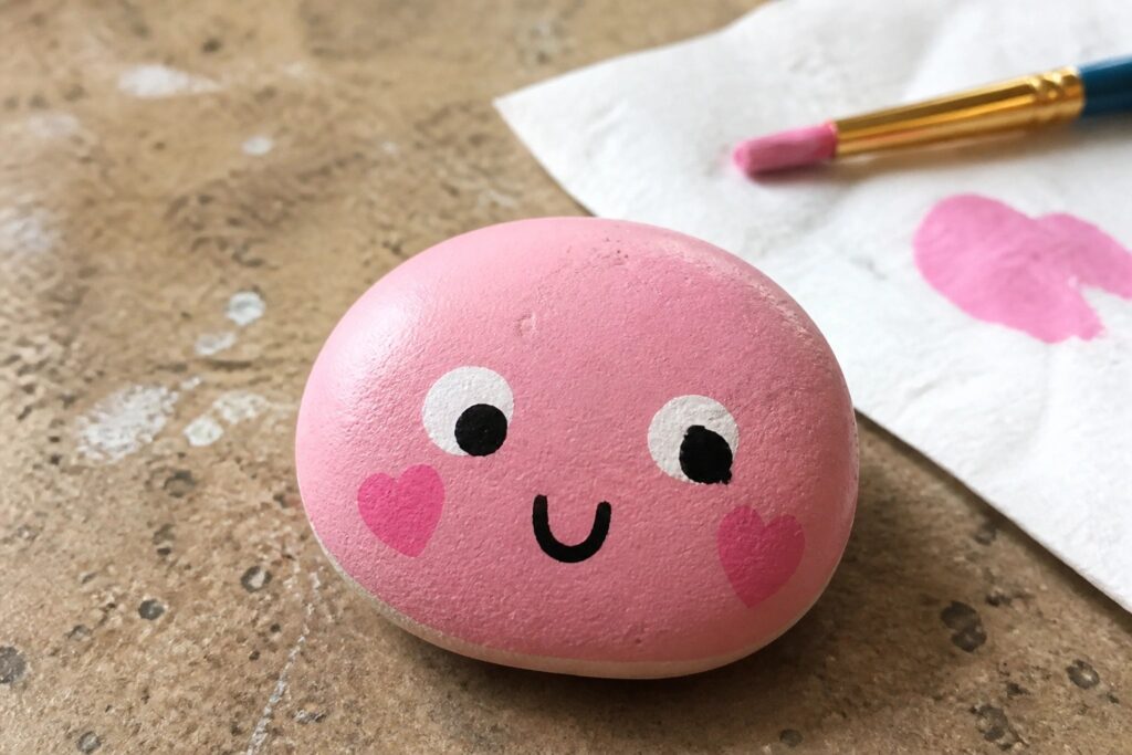 Pink heart-shaped painted rock with cute face beside paintbrush and towel