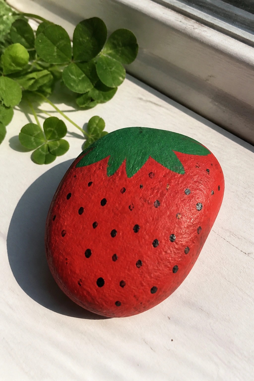 Painted red strawberry rock with green top on white windowsill near leaves